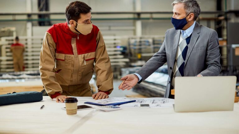 A factory worker and a manager wearing face masks discuss paper blueprints on a table in a manufacturing facility.