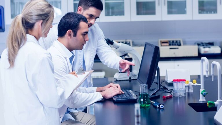 Hospital laboratory staff evaluating compliance and reliability data on a computer system, with a microscope and lab equipment in the background.