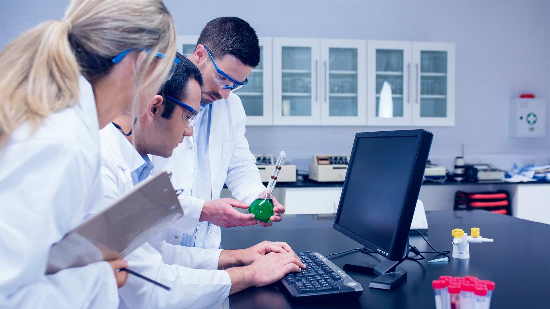 Three lab researchers in protective gear inspecting a chemical sample while entering data on a computer.