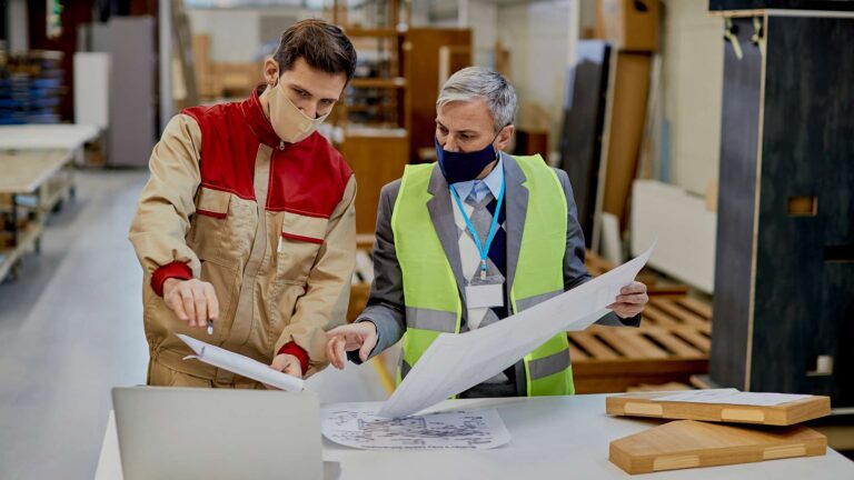 A manufacturing supervisor and technician wearing face masks review digital software on a laptop and physical blueprints to inspect wooden components against design standards on the factory floor.