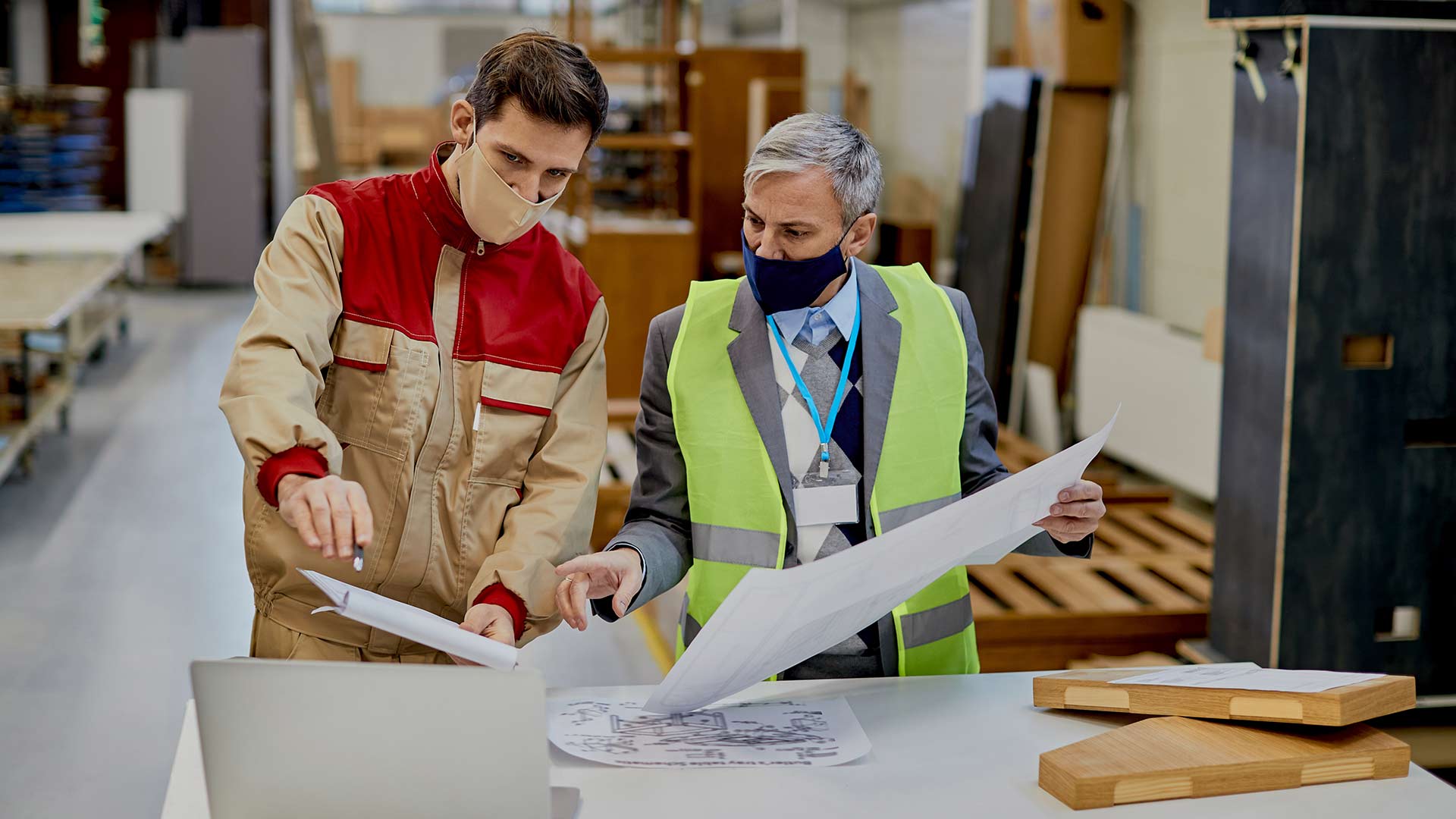 A manufacturing supervisor and technician wearing face masks review digital software on a laptop and physical blueprints to inspect wooden components against design standards on the factory floor.
