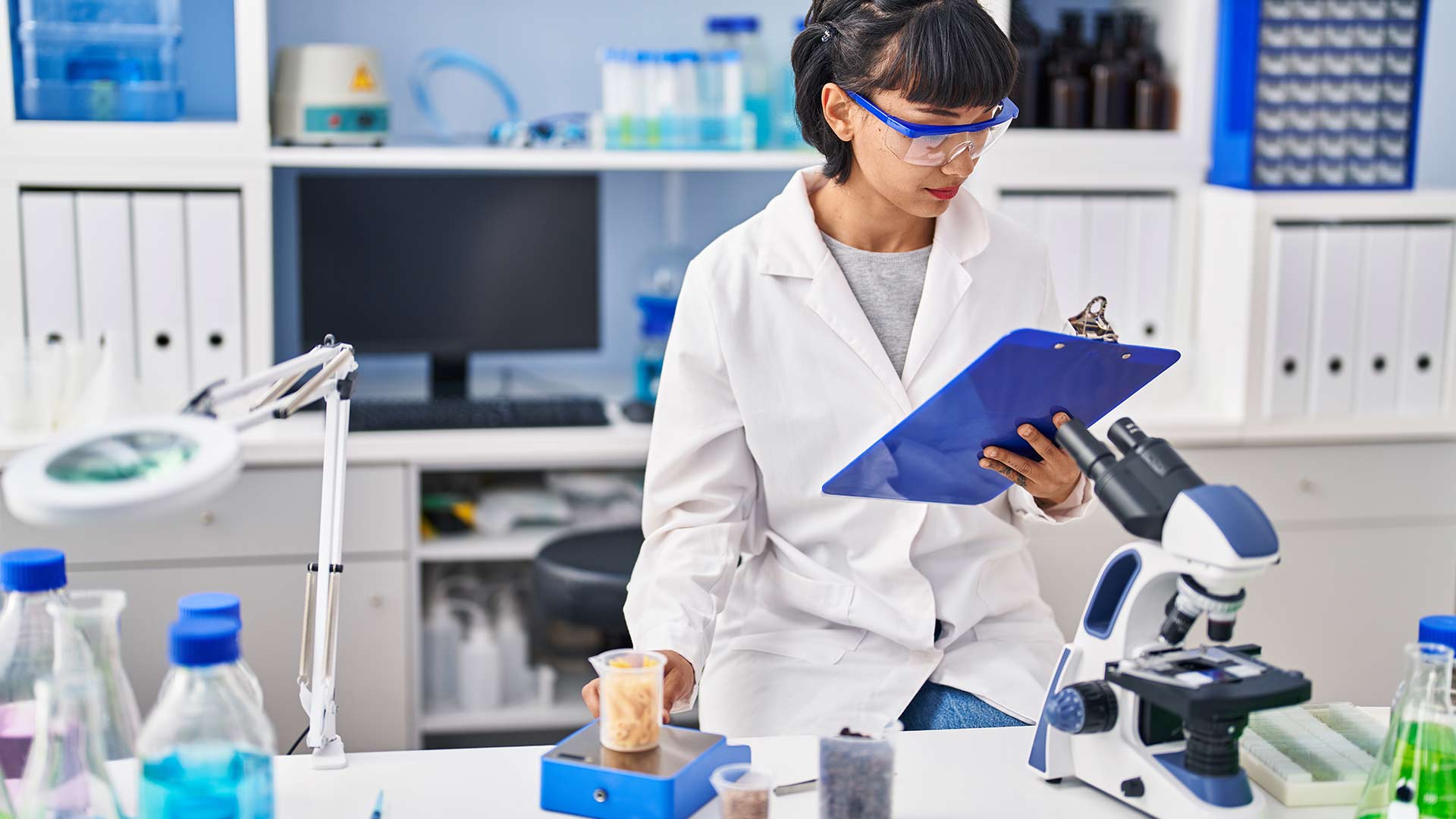 A female lab technician in a lab coat writing on a clipboard while examining lab samples next to a microscope.