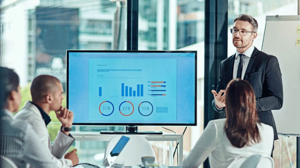 Business professional standing in a modern boardroom, pointing to data charts and graphs on a display screen while presenting to colleagues seated around a table.