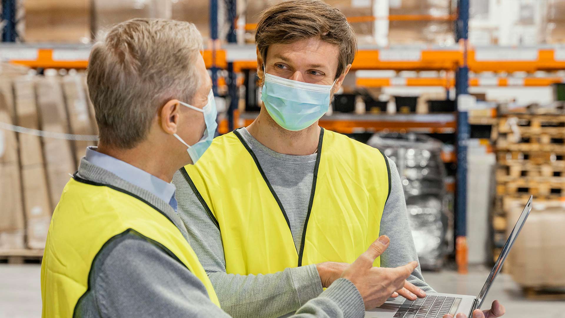Two male quality assurance inspectors wearing face masks and safety vests discussing data on a laptop in a manufacturing warehouse.