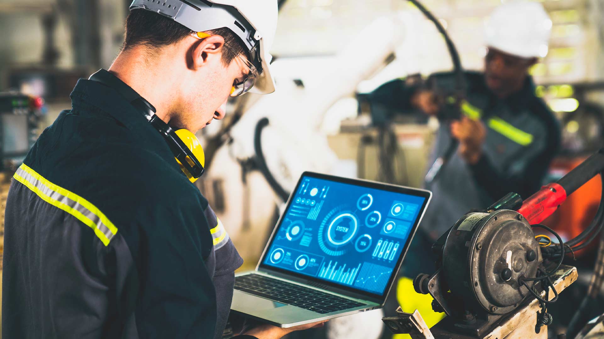 An engineer wearing safety gear reviewing equipment status and maintenance data on a laptop dashboard inside an industrial setting.