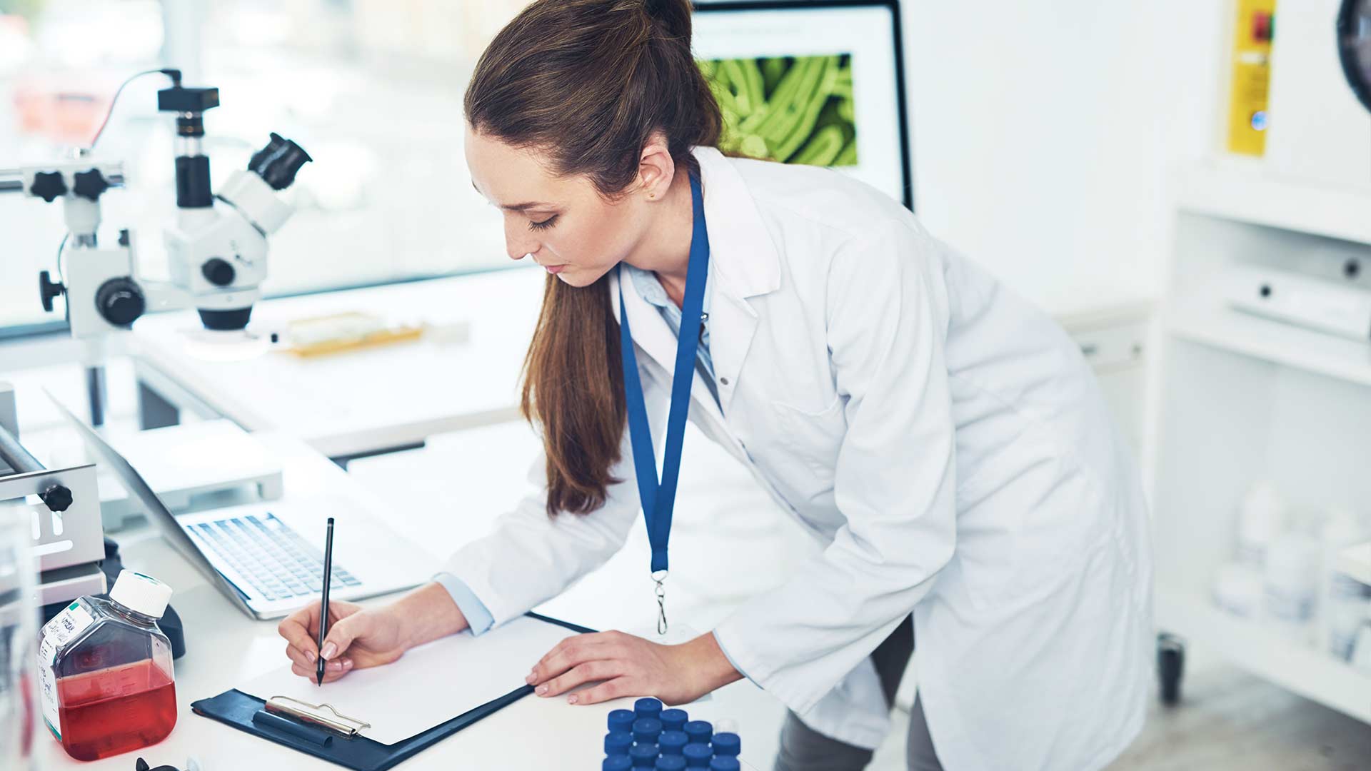 A female scientist in a lab coat writing notes on a clipboard next to sample bottles, a microscope, and a laptop for audit compliance and sample tracking.